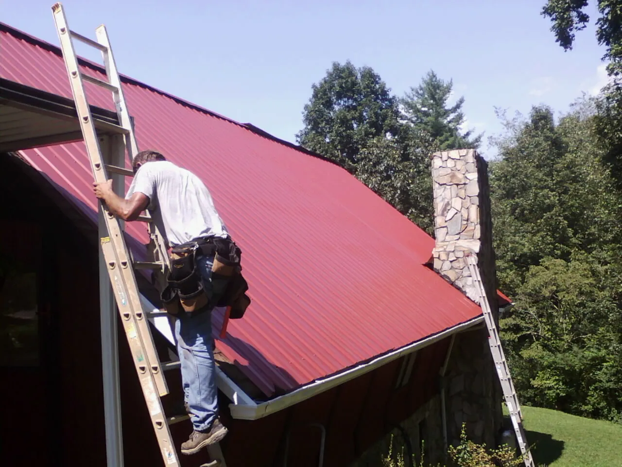Roofer on a ladder beside a red metal roof and stone chimney — Soffit & Fascia Repair by Roofing Brothers in Silver Plume